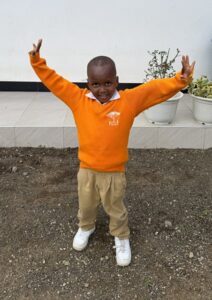 Image of young school age boy smiling with arms raised in oragne sweater and khaki pants